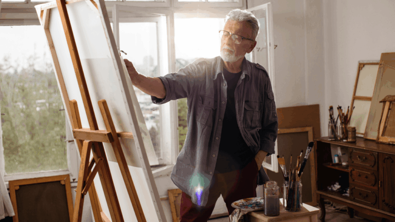 An older man stands in front of an easel, painting in a sunny room.